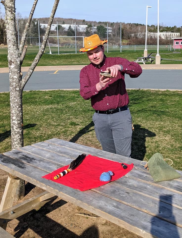Shawn Feener standing outside beside a tree and picnic table. he holds plants and a shell that will be used in the smudge..