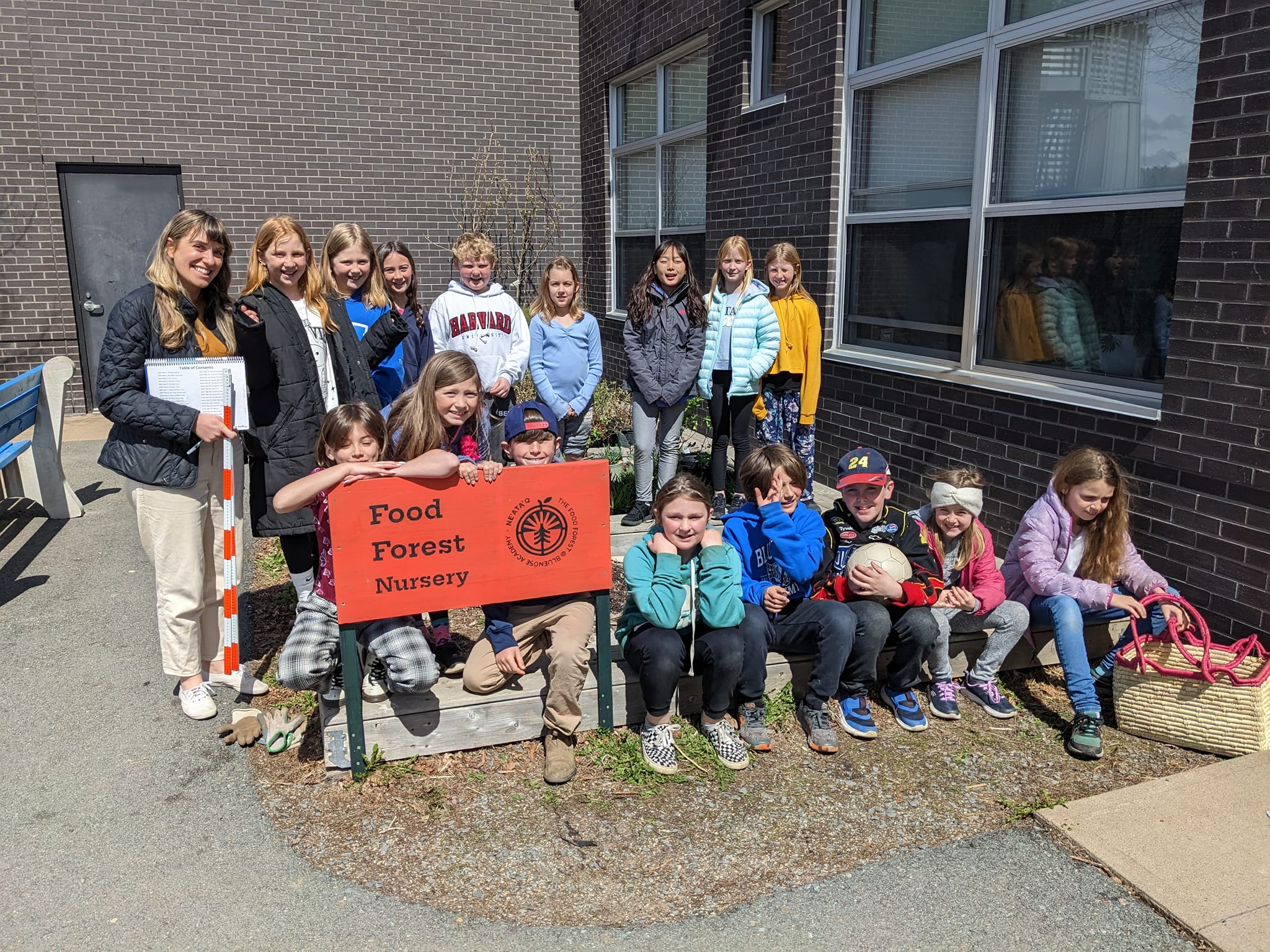 Group photo of a grade 3/4 class sitting and standing outside near the school and the food forest nursery.