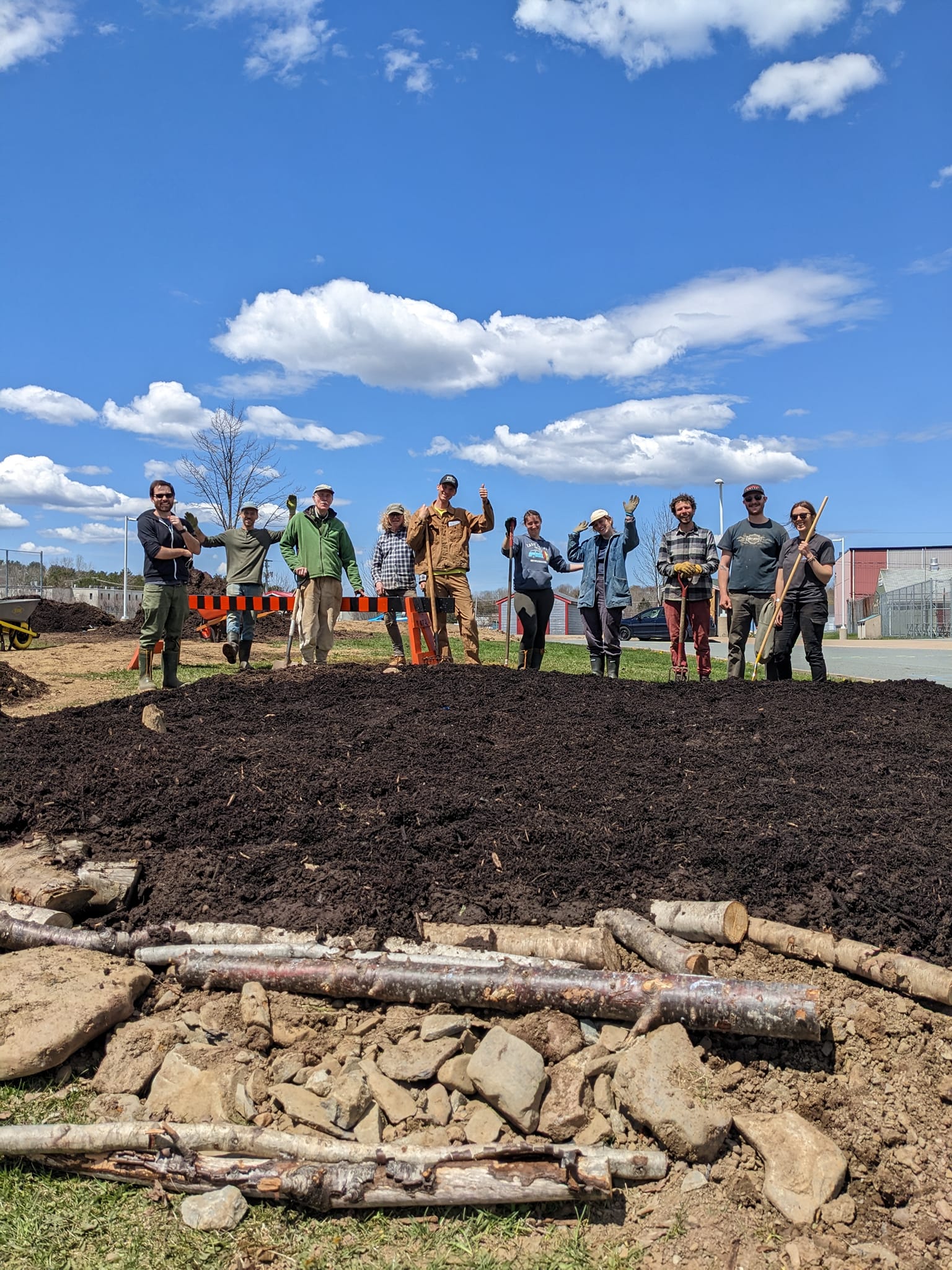 Ten people in a line holding shovels and/or waving. In the foreground, the ground is prepared for planting and covered with mulch.